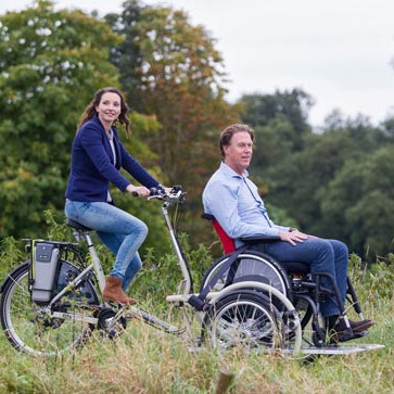 E-Rollstuhlfahrrad VeloPlus von Van Raam auf einem Feldweg. Vorne sitzt ein Rollstuhlfahrer im Rollstuhl und hinten tritt eine Frau in die Pedale.